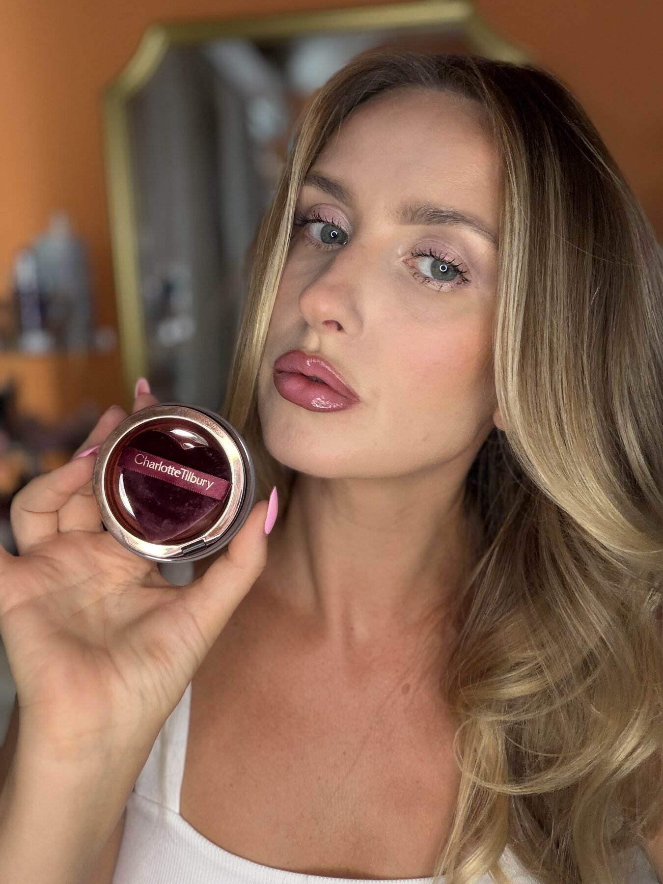 Woman puckers her glossy lips while holding a heart-shaped Charlotte Tilbury lip product compact near a mirror.