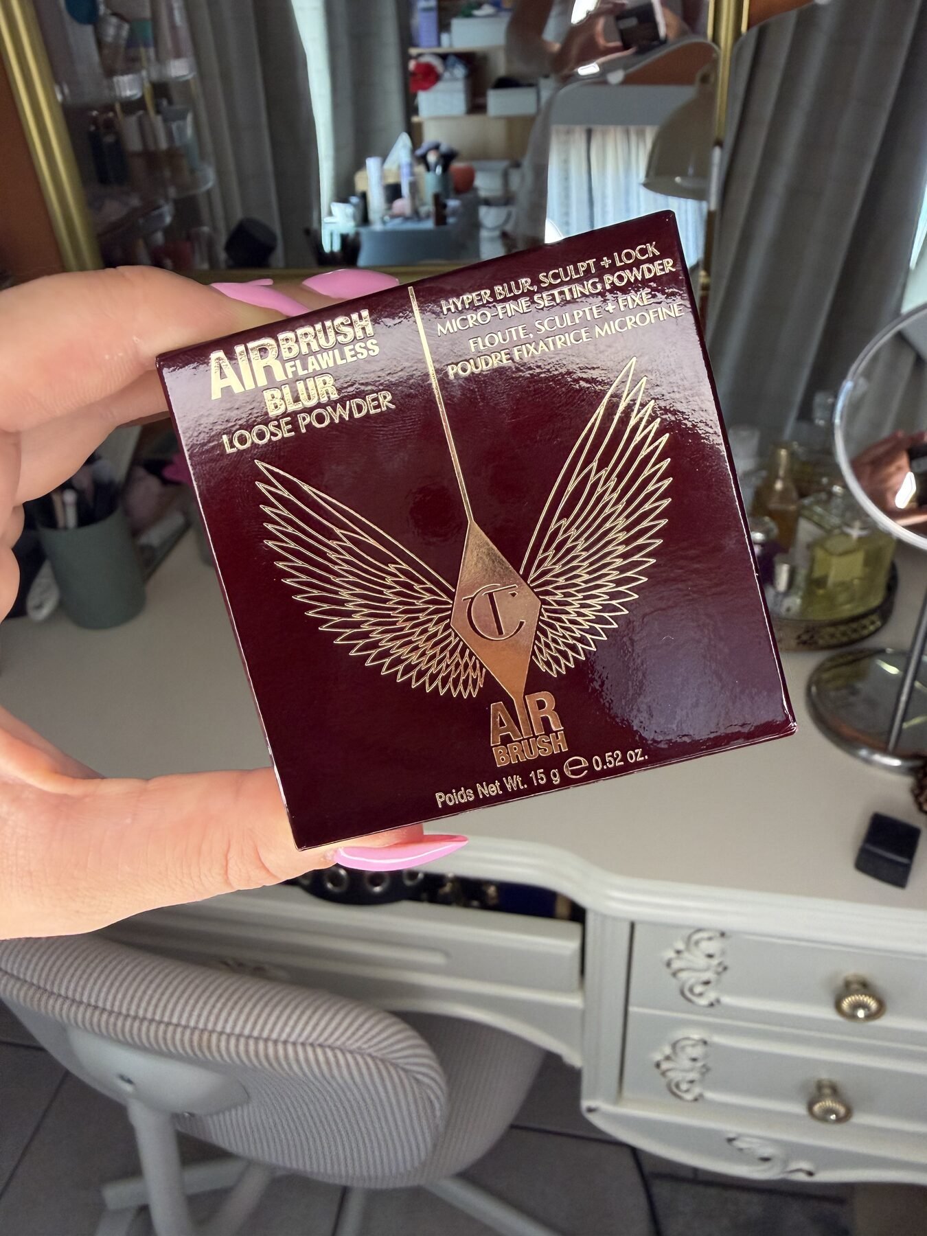 Hand with pink nails holds Charlotte Tilbury Airbrush Flawless Blur Loose Powder box at a vanity table.