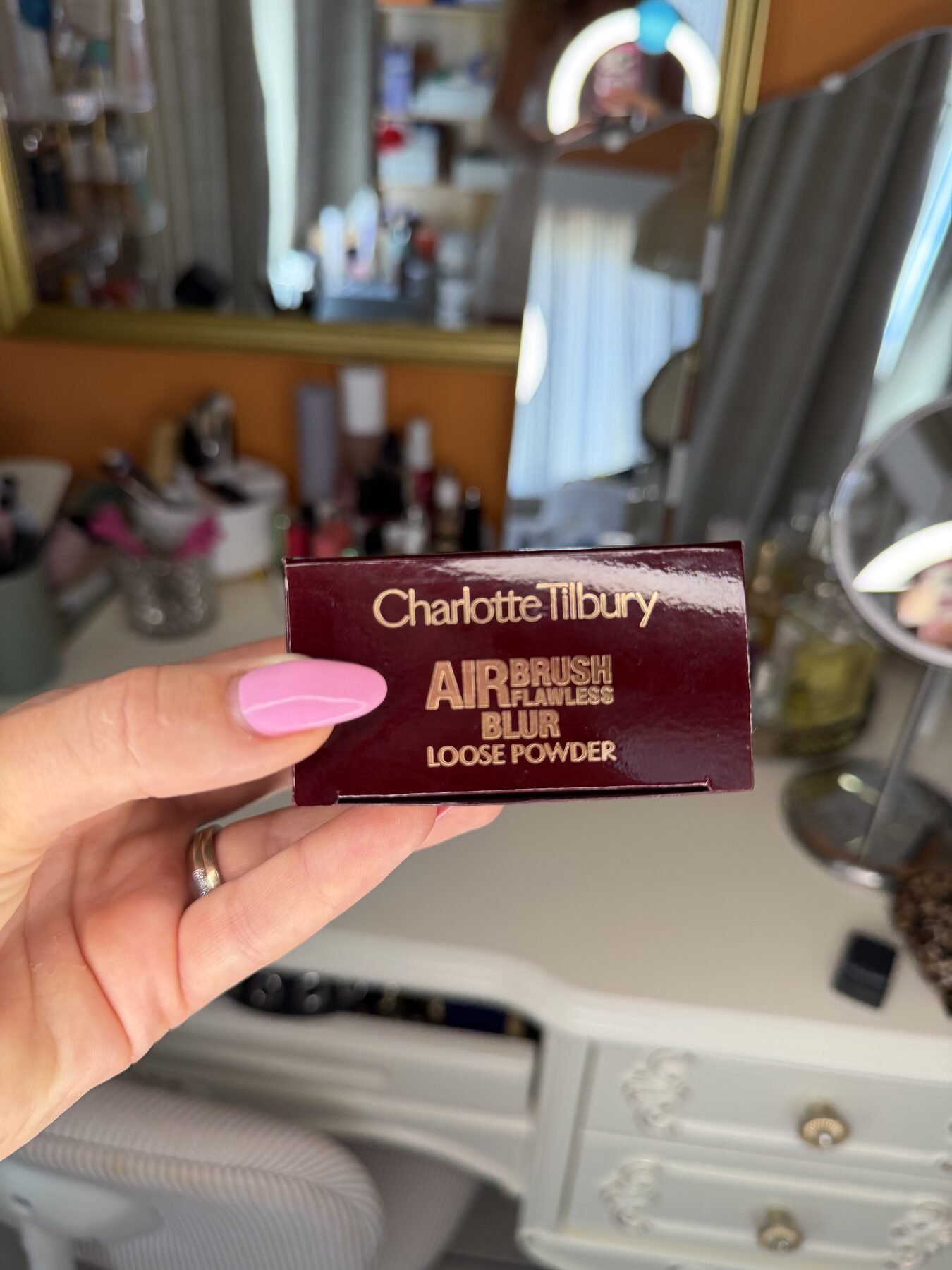 Hand with pink manicure holds Charlotte Tilbury Airbrush Flawless Blur Loose Powder box at a mirrored vanity.