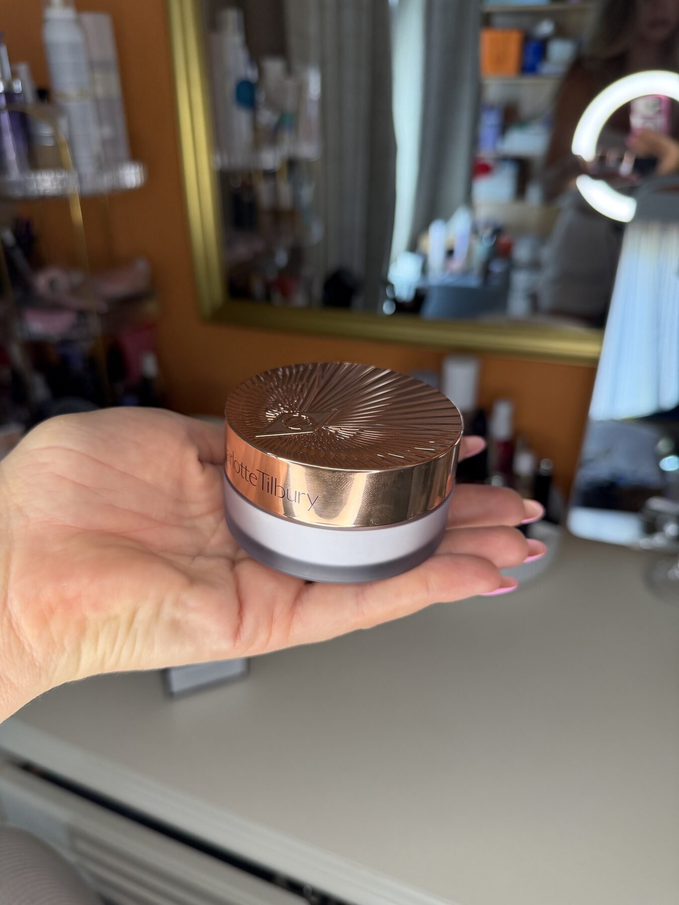 Hand holding a Charlotte Tilbury loose powder jar with gold lid at a mirrored vanity.