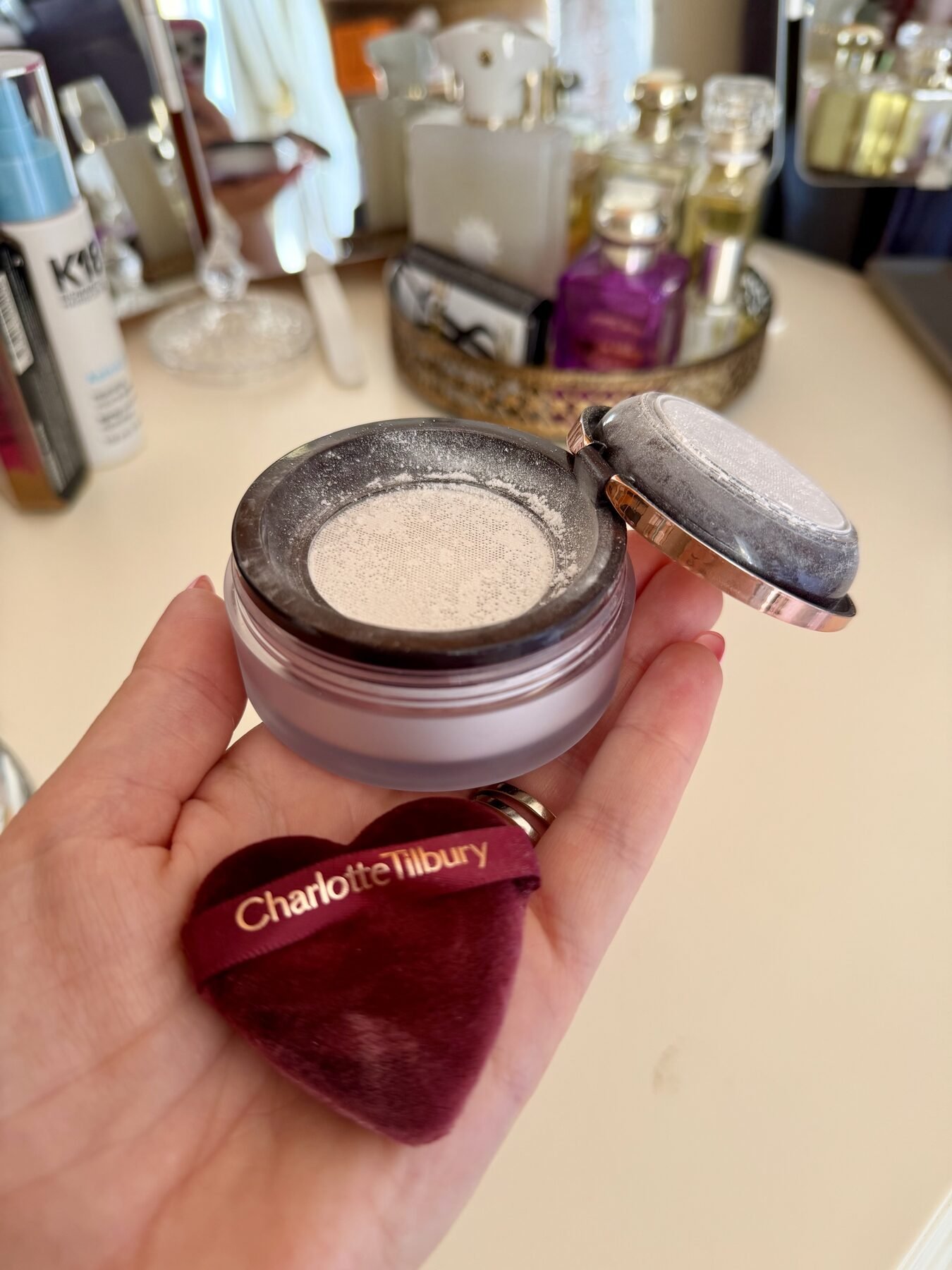 Hand holding open Charlotte Tilbury setting powder jar and heart-shaped puff on a vanity table.