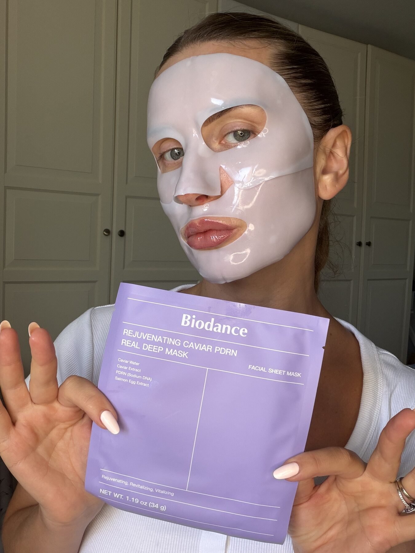 Woman wearing a white sheet mask holds up its purple Biodance Rejuvenating Caviar PDRN packaging.