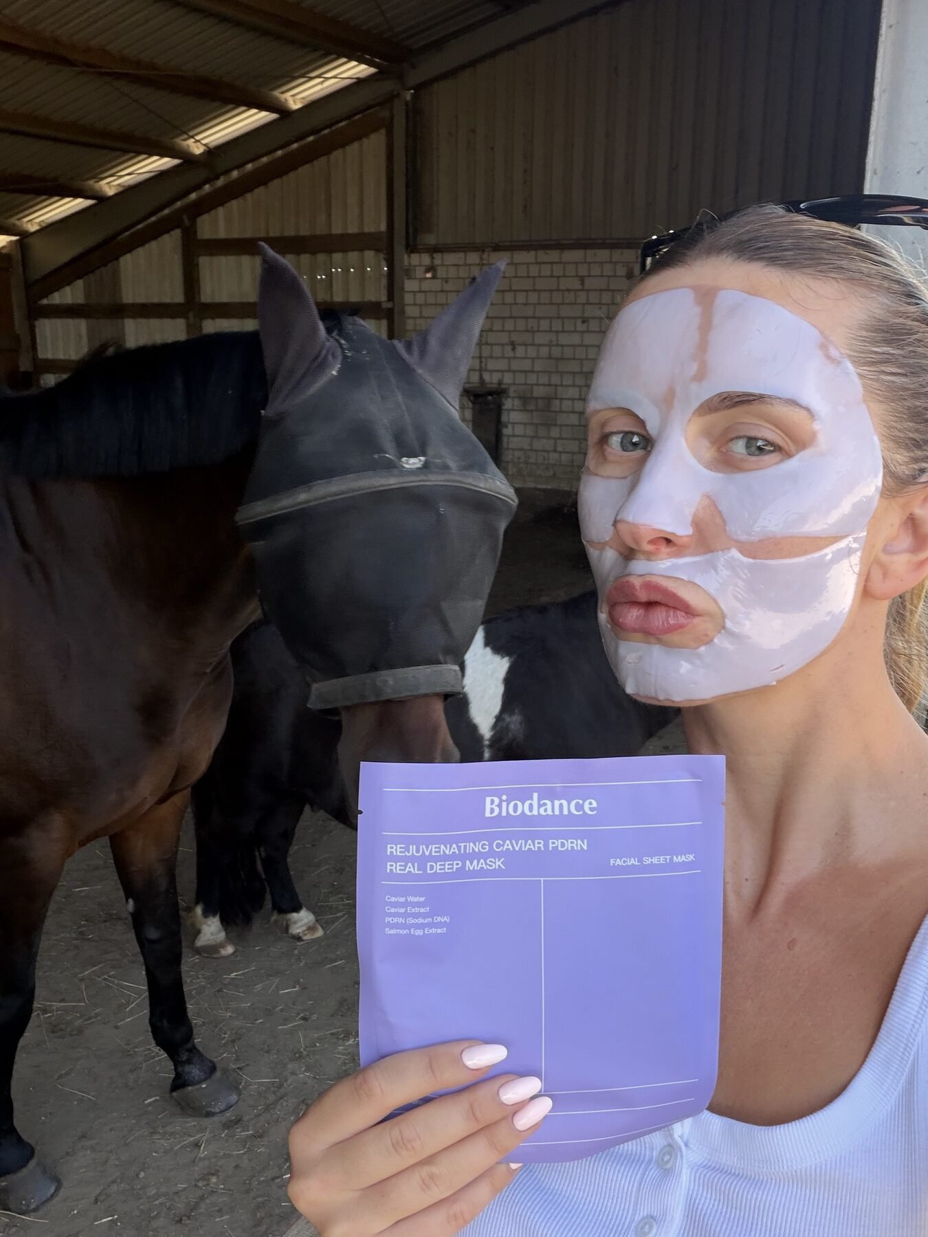Woman wearing a Biodance sheet mask holds the packaging near horses in a stable.