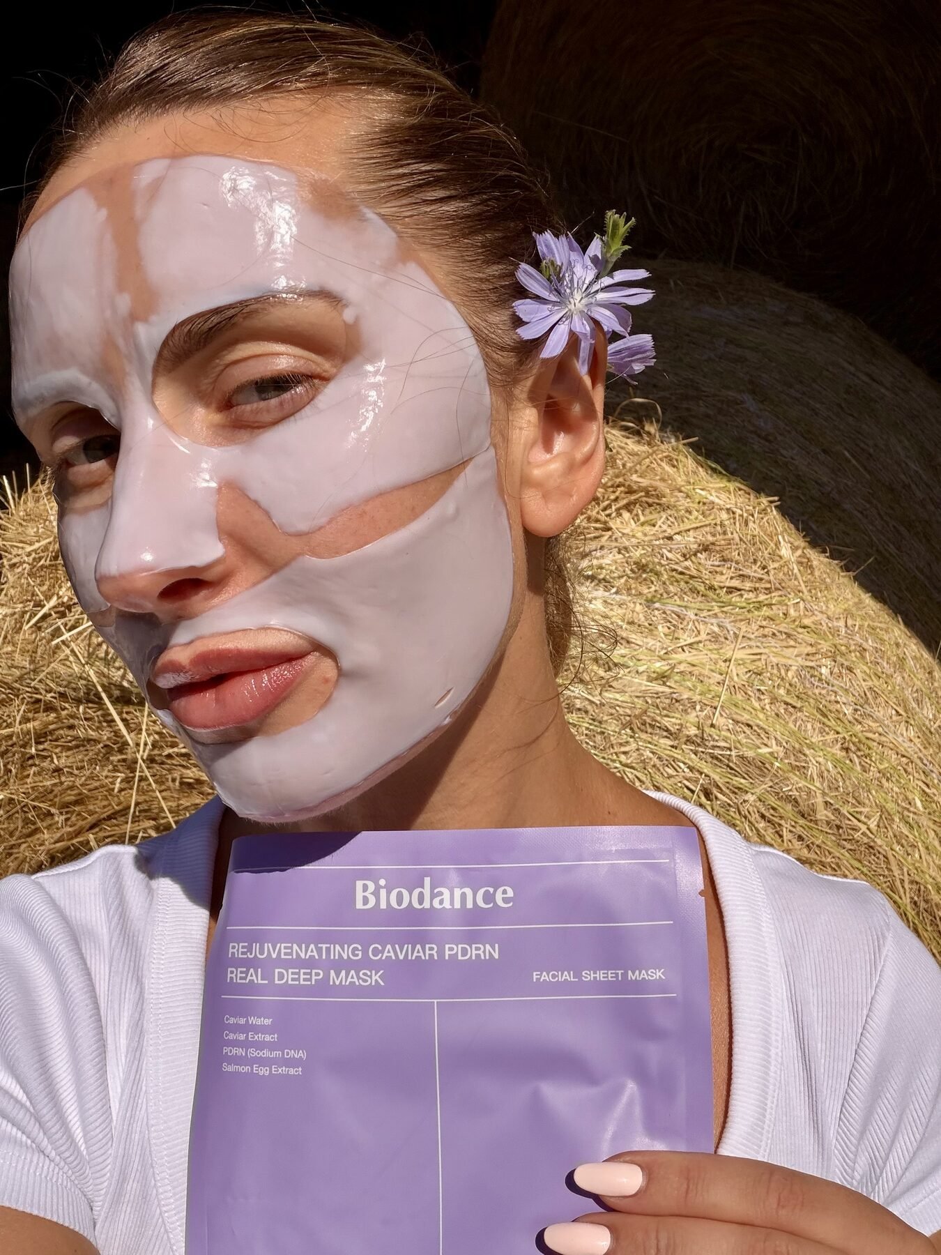 Woman wearing a Biodance sheet mask holds the product package while sitting near hay bales outdoors.