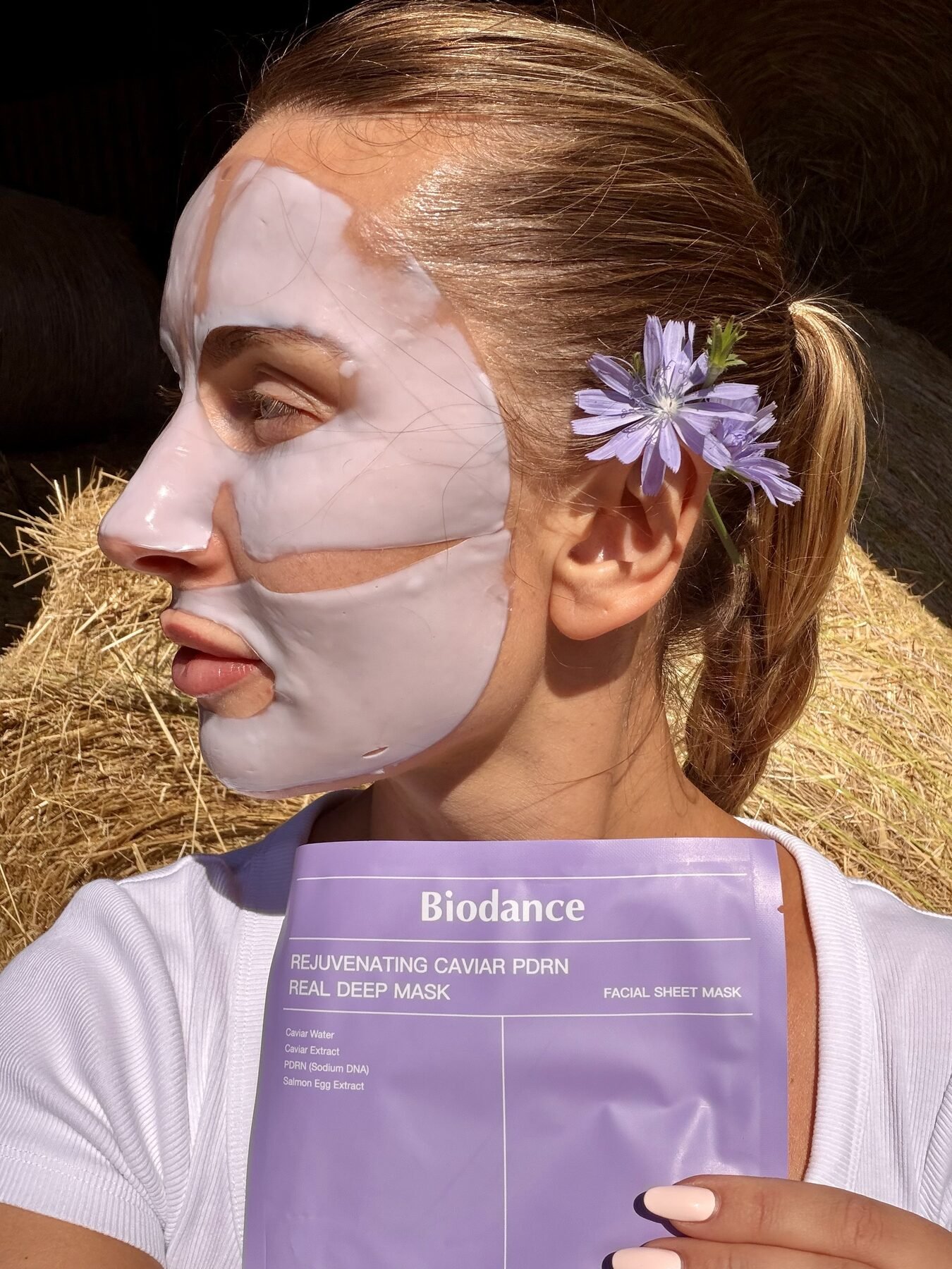 Woman wearing a Biodance sheet mask with a purple flower in her hair, holding the product packaging outdoors.