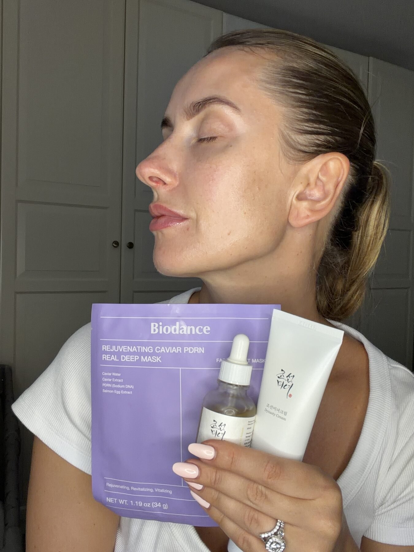 Woman in profile with eyes closed holds a Biodance sheet mask, serum, and cream.