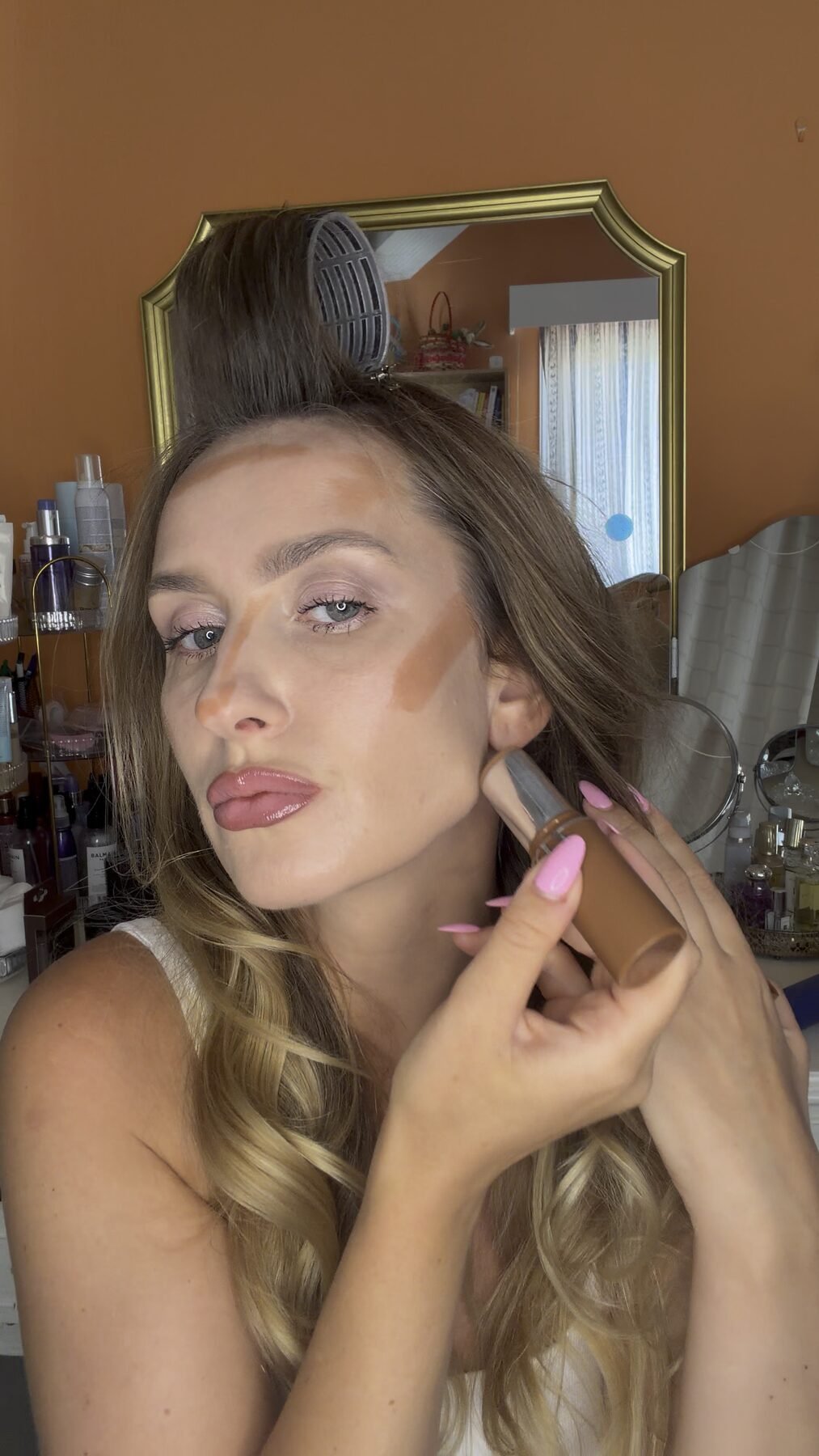 Woman holding bronzer tube, applying contour stripes across forehead, nose, and cheeks at makeup vanity.