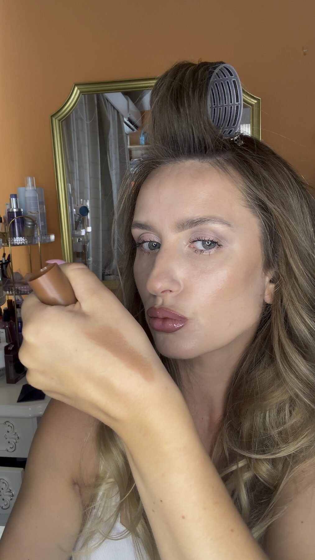 Woman with a hair roller swatches Dior bronzer on her hand at a vanity mirror.