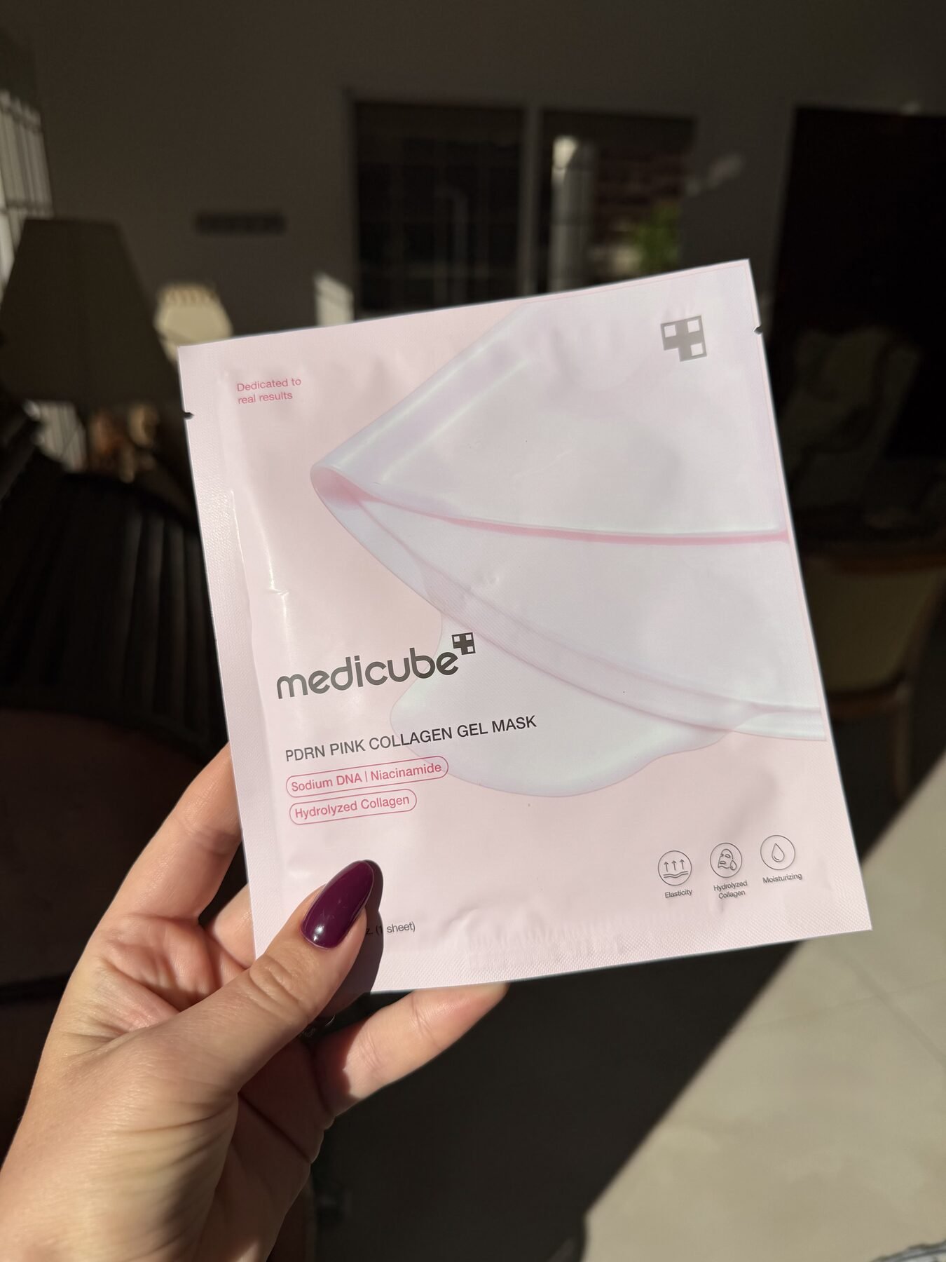 Hand with dark purple nail polish holds a Medicube PDRN Pink Collagen Gel Mask sachet indoors.
