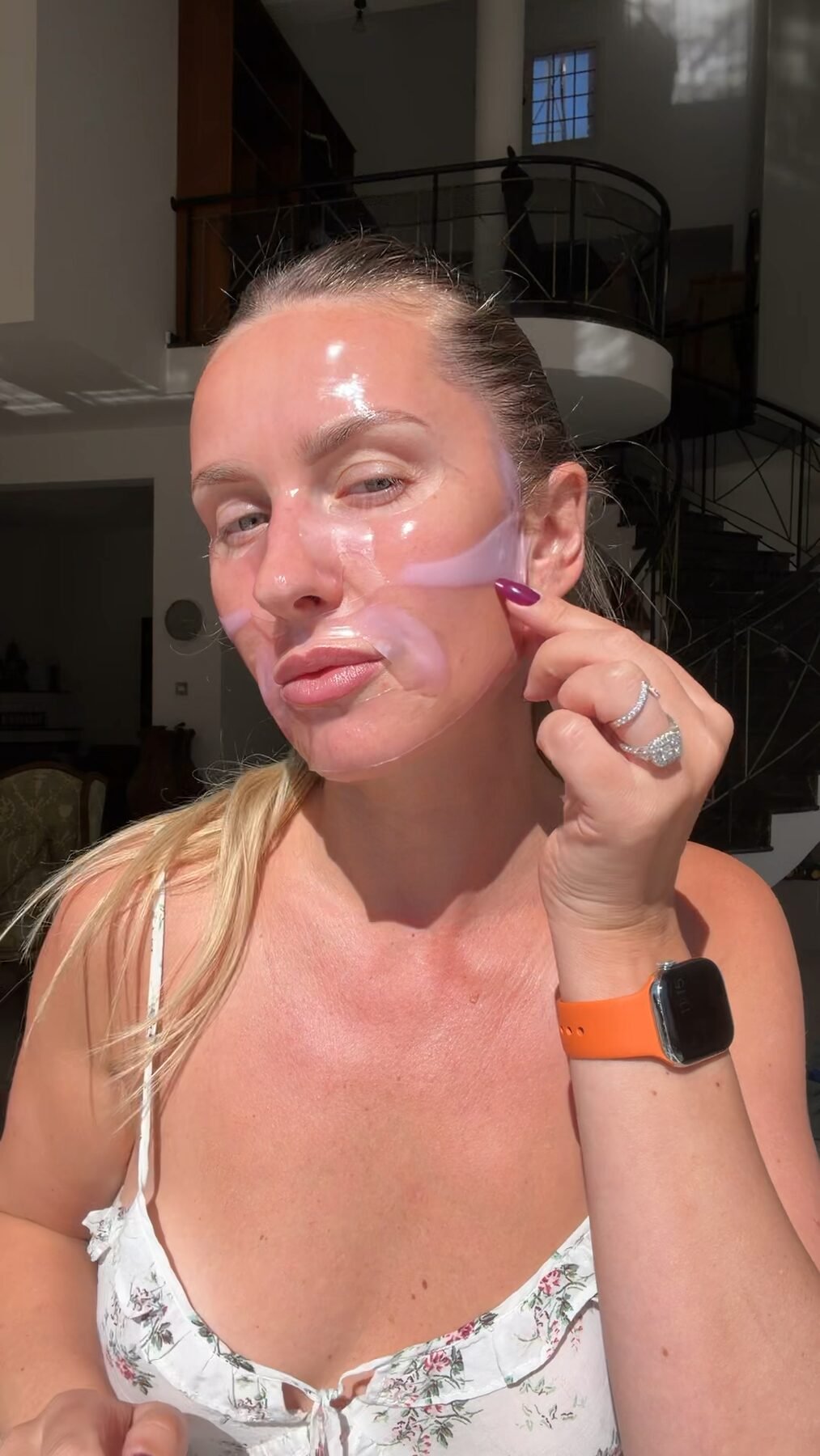 Woman applies a clear hydrating gel sheet mask to her face in a sunlit, multi-level living room.