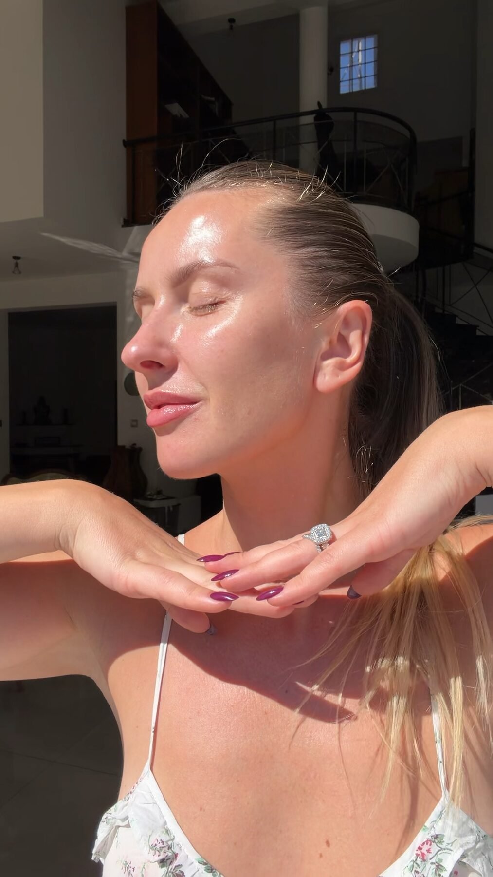 Woman with glowing, dewy skin tilts her head back, eyes closed, hands raised near her ponytail in sunlight.
