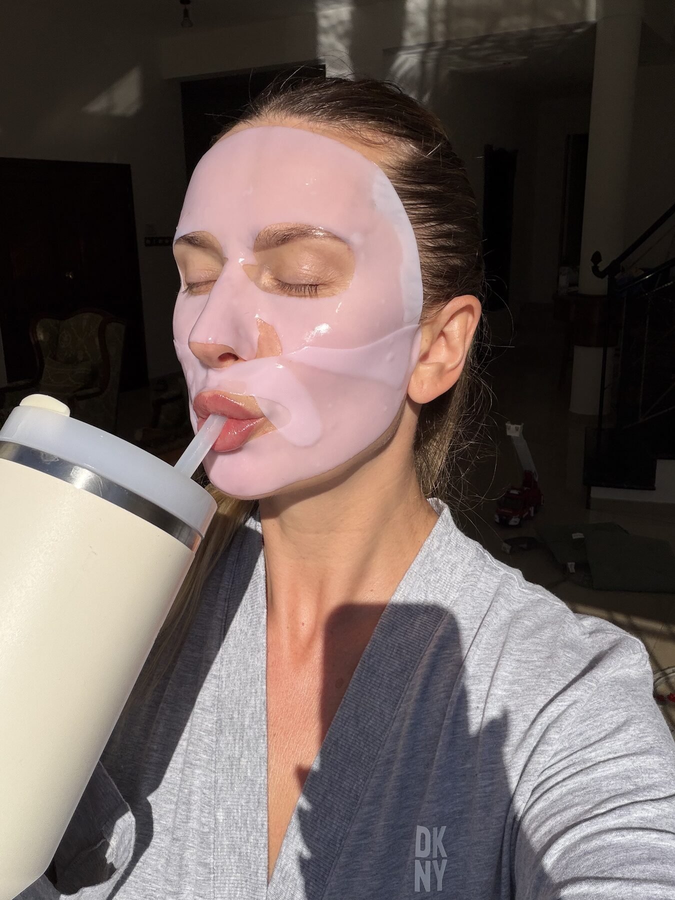 Woman wearing a pink sheet mask sips from a tumbler with a straw indoors.