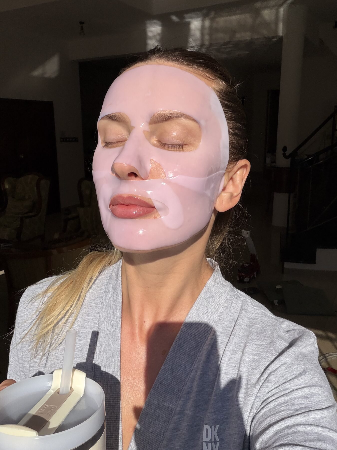 Woman with eyes closed wears a pink sheet mask on her face, holding a mixing cup indoors.