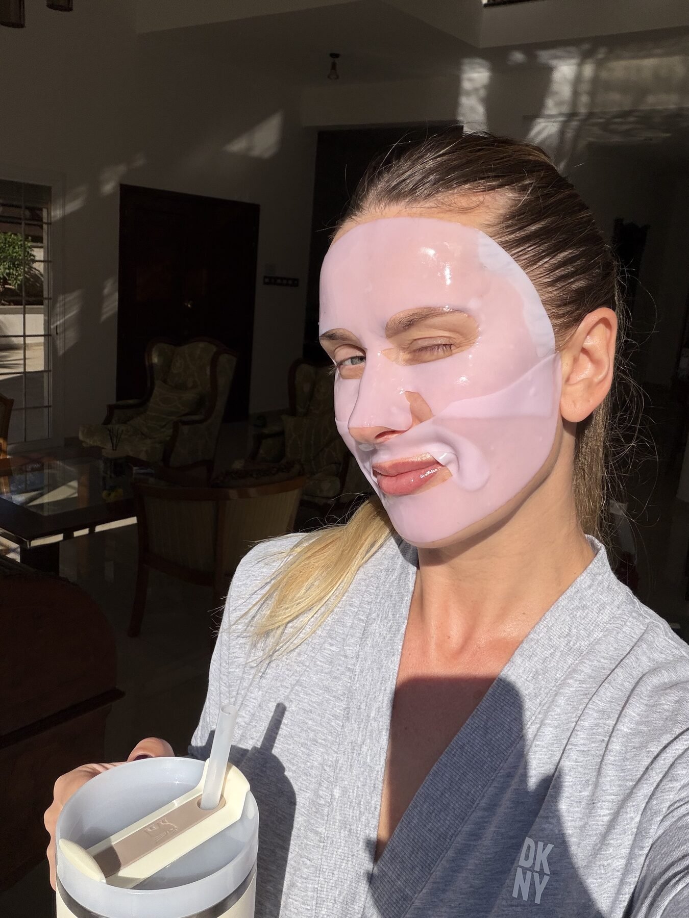 Woman wearing a pink sheet mask winks while holding a tumbler, standing in a sunlit living room.