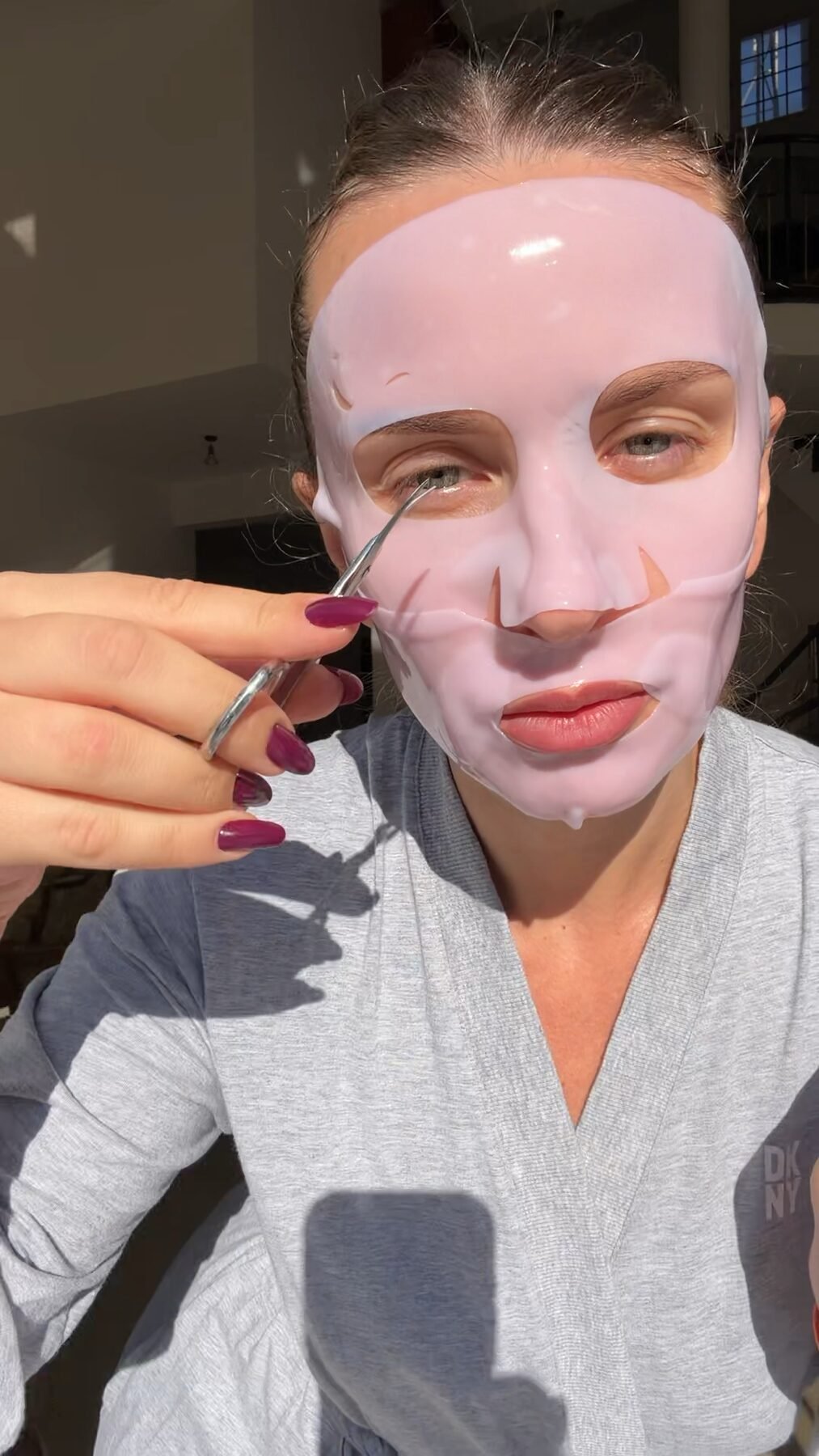 Woman wears a pink gel sheet mask, adjusting it near her eye with a small tool, taking a selfie indoors.
