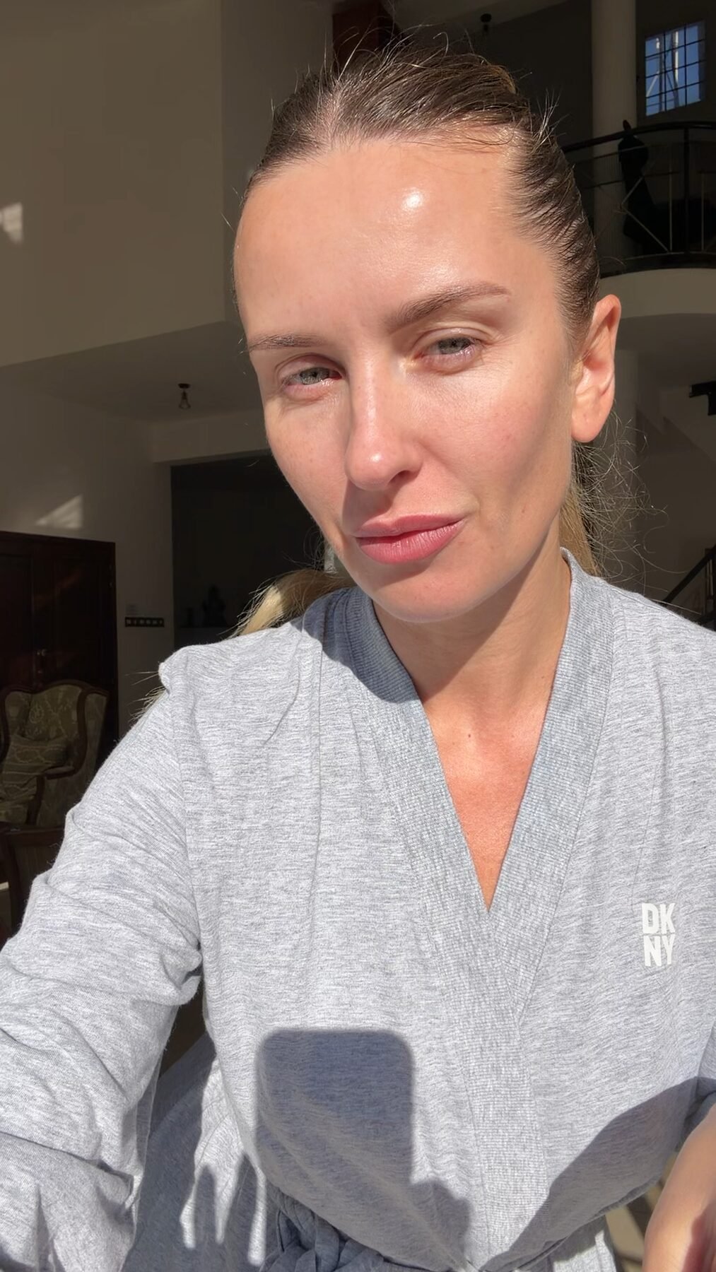 Woman with damp hair and bare, dewy skin takes a selfie in a bright, high-ceilinged home.