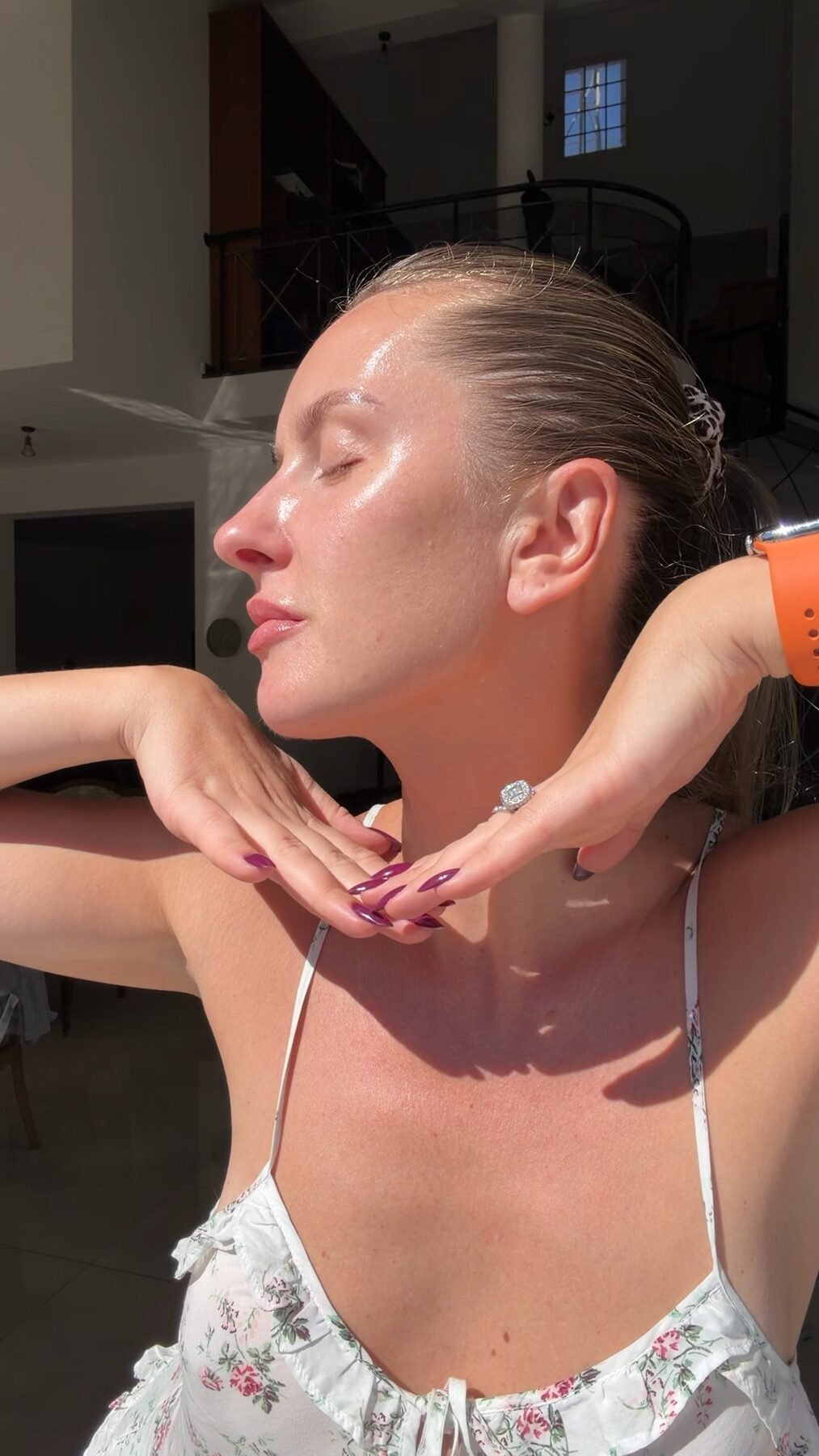Woman with glossy, dewy skin tilts her head back, eyes closed, hands behind her neck indoors.