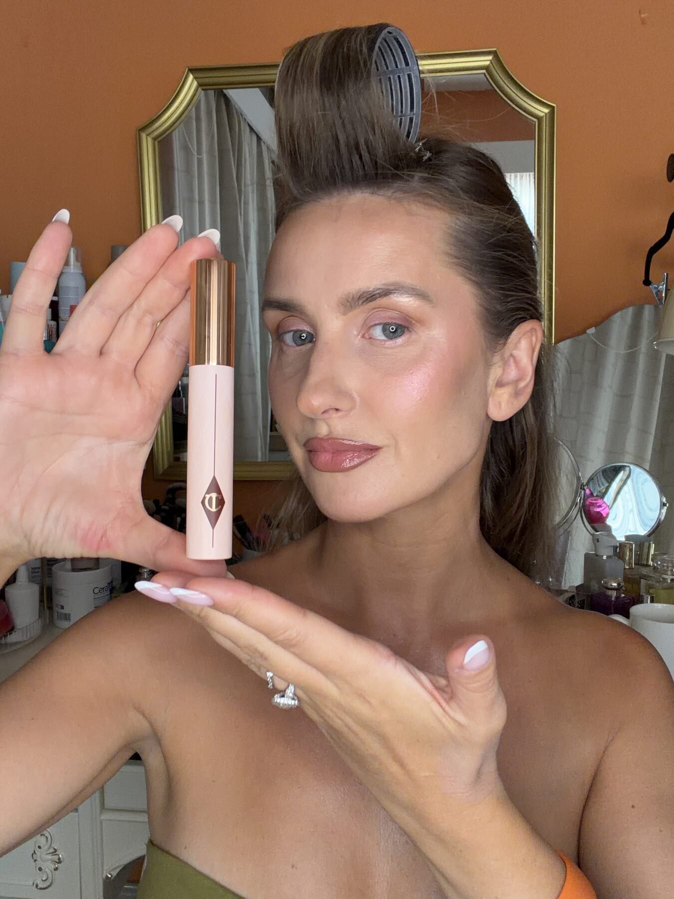 Woman with hair in a roller holds up a Charlotte Tilbury mascara tube in her bathroom.