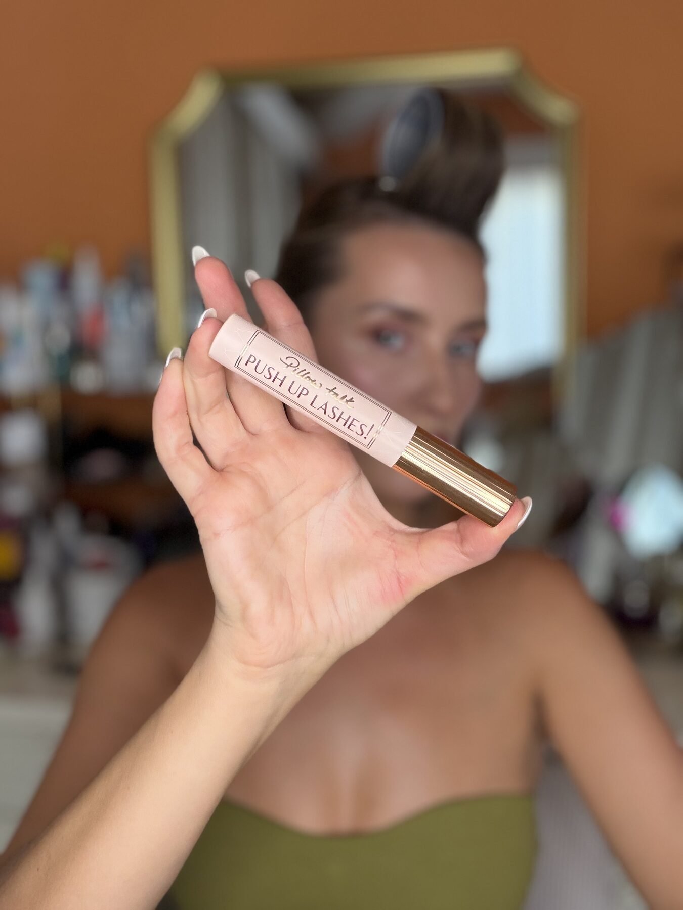 Woman holds up Charlotte Tilbury Pillow Talk Push Up Lashes mascara tube in front of bathroom mirror.