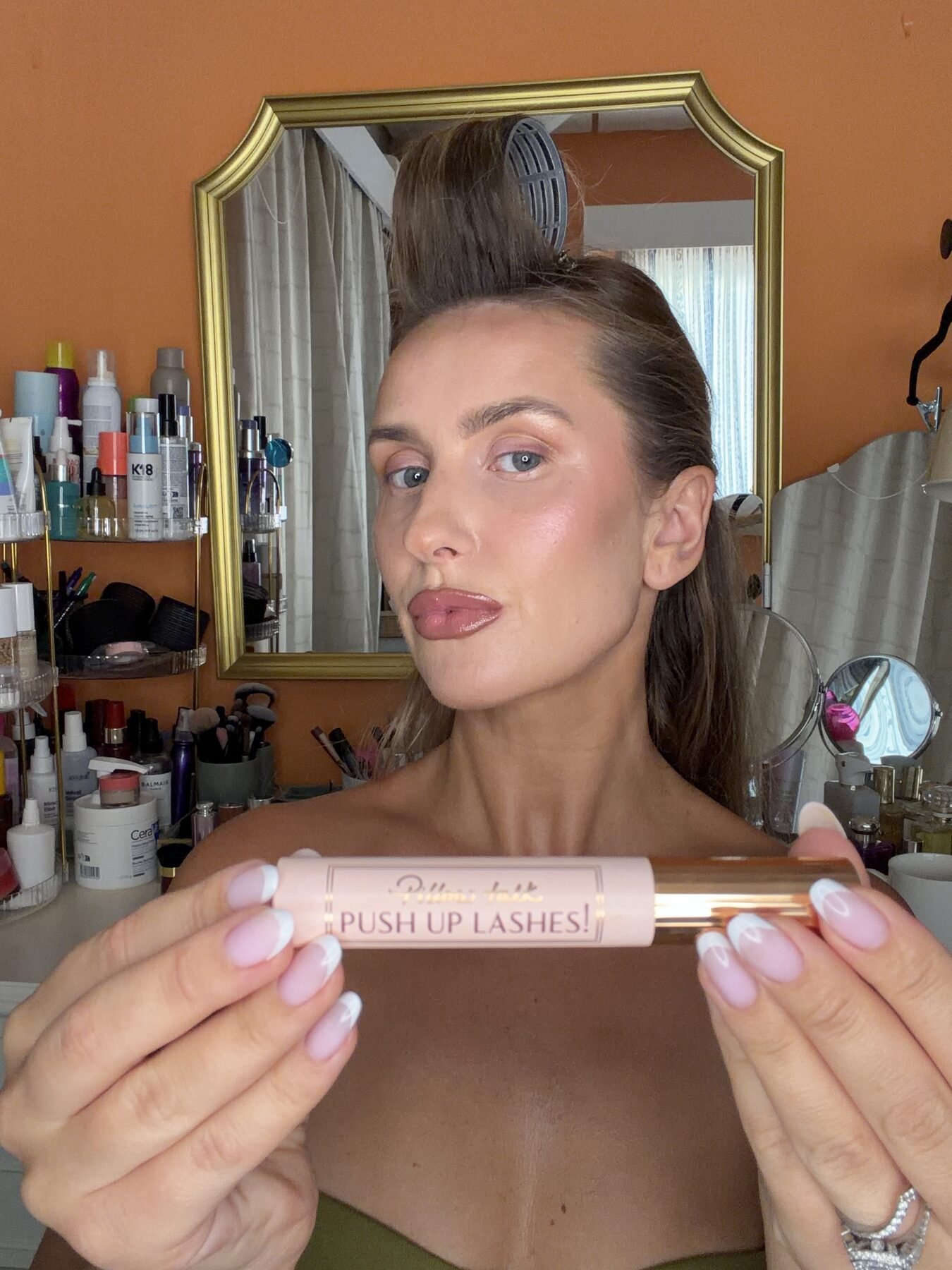 Woman with hair clipped up holds a Charlotte Tilbury Pillow Talk Push Up Lashes mascara near a vanity mirror.