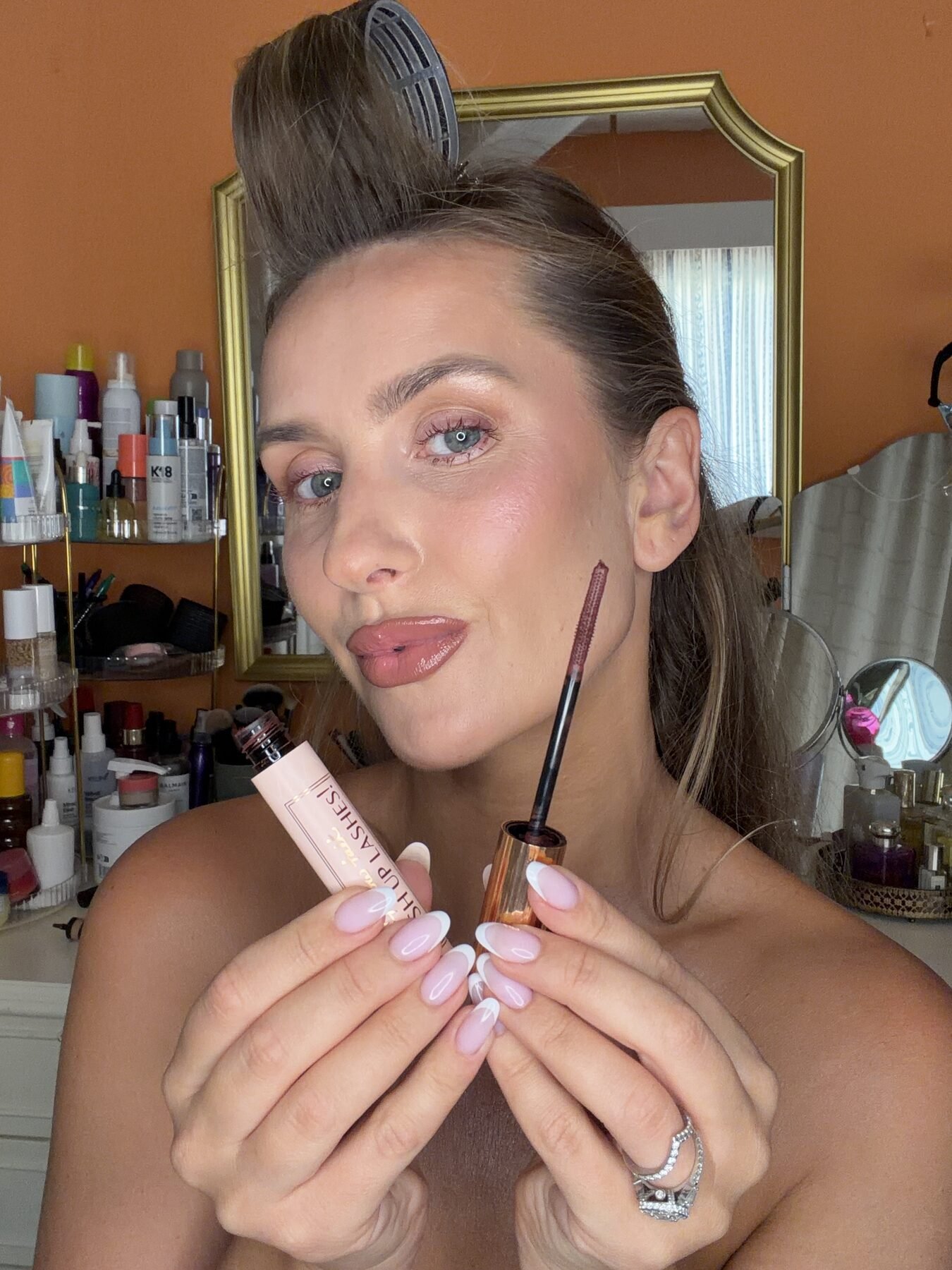 Woman with hair clipped up holds an open mascara wand and its tube near her face in a bathroom.