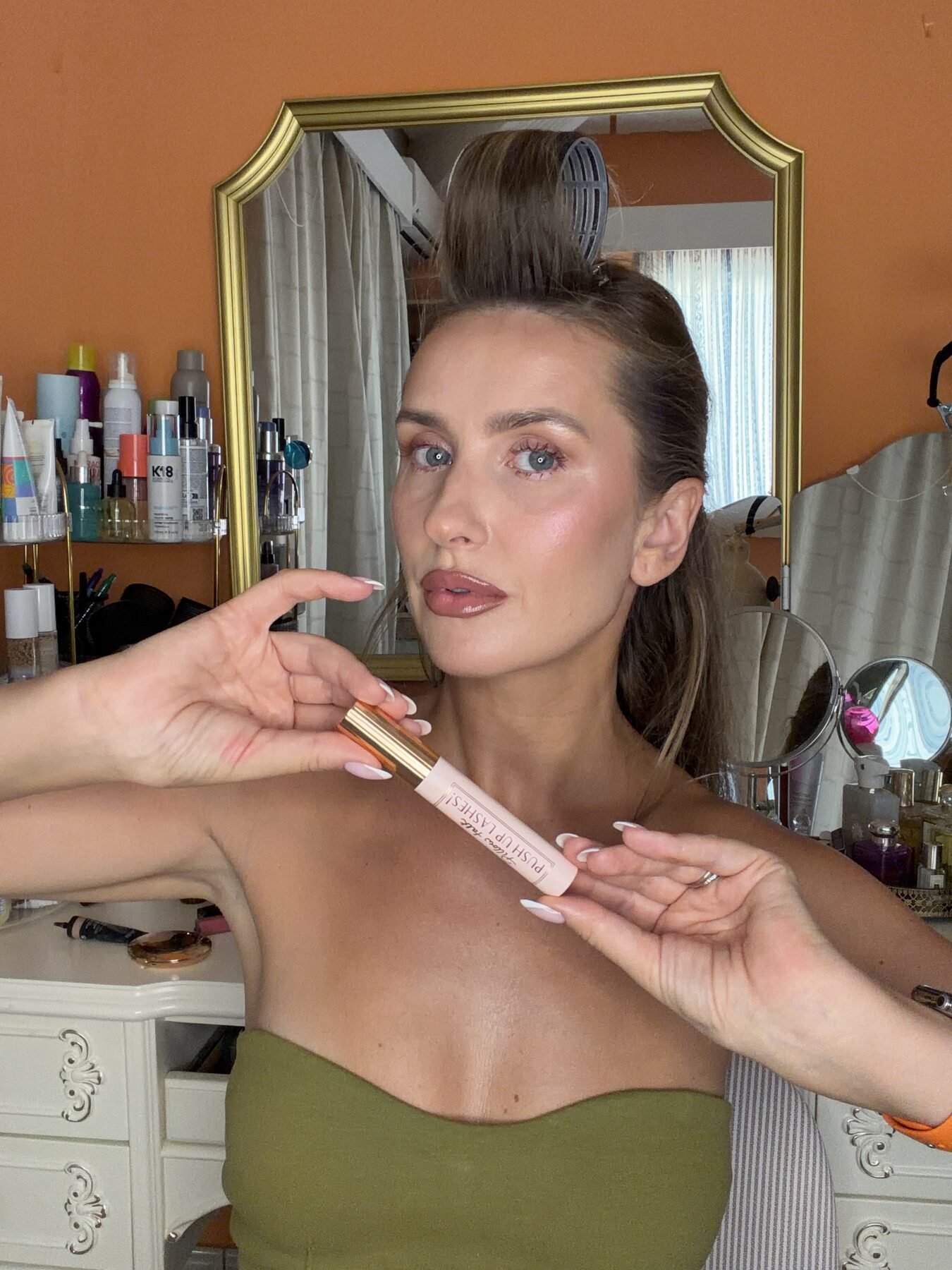 Woman with hair rollers holds up a Charlotte Tilbury mascara tube at her vanity mirror.