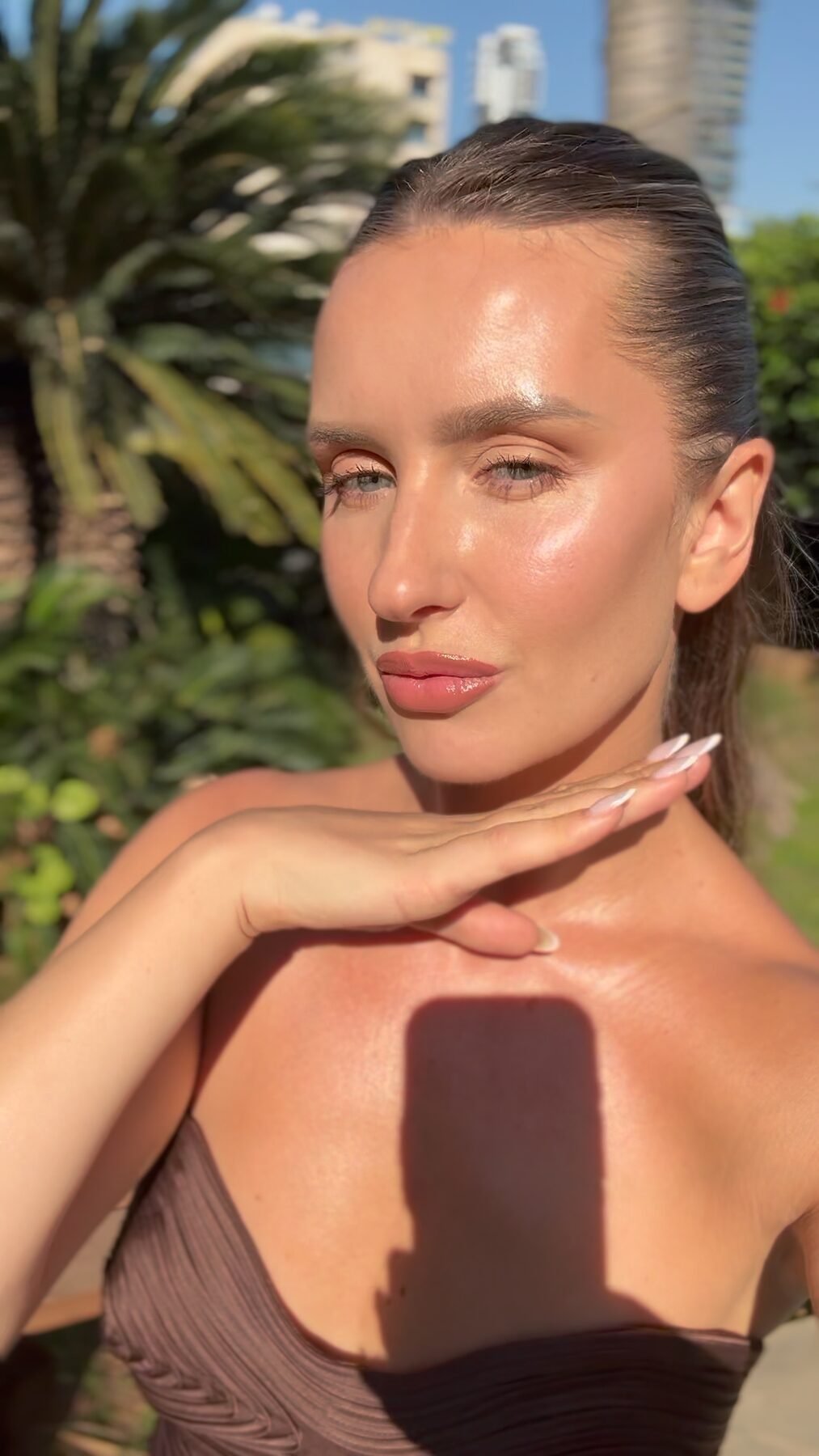 Close-up selfie of a woman with dewy, glowing skin outdoors, phone shadow visible on her chest.