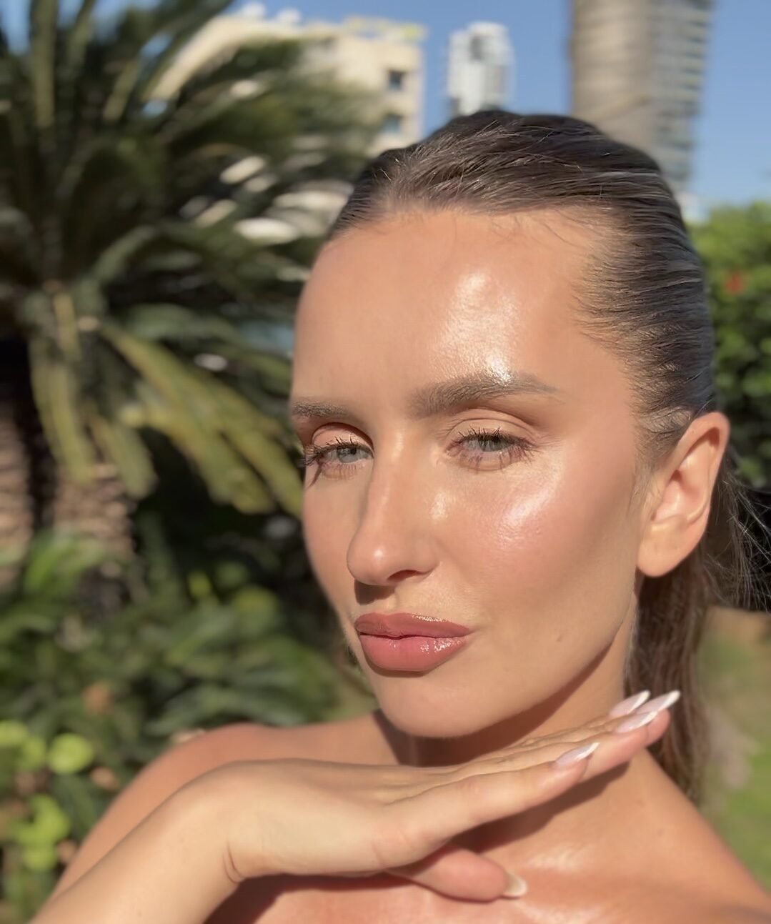 Close-up of a woman with glowing, dewy makeup and slicked-back hair, hand near her chin outdoors.