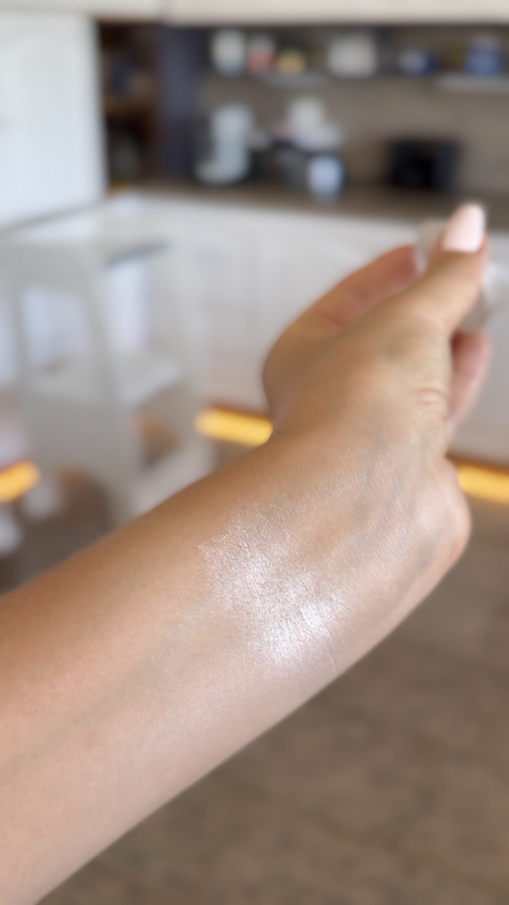 Close-up of a shimmery highlighter swatch on the back of a hand in a kitchen.