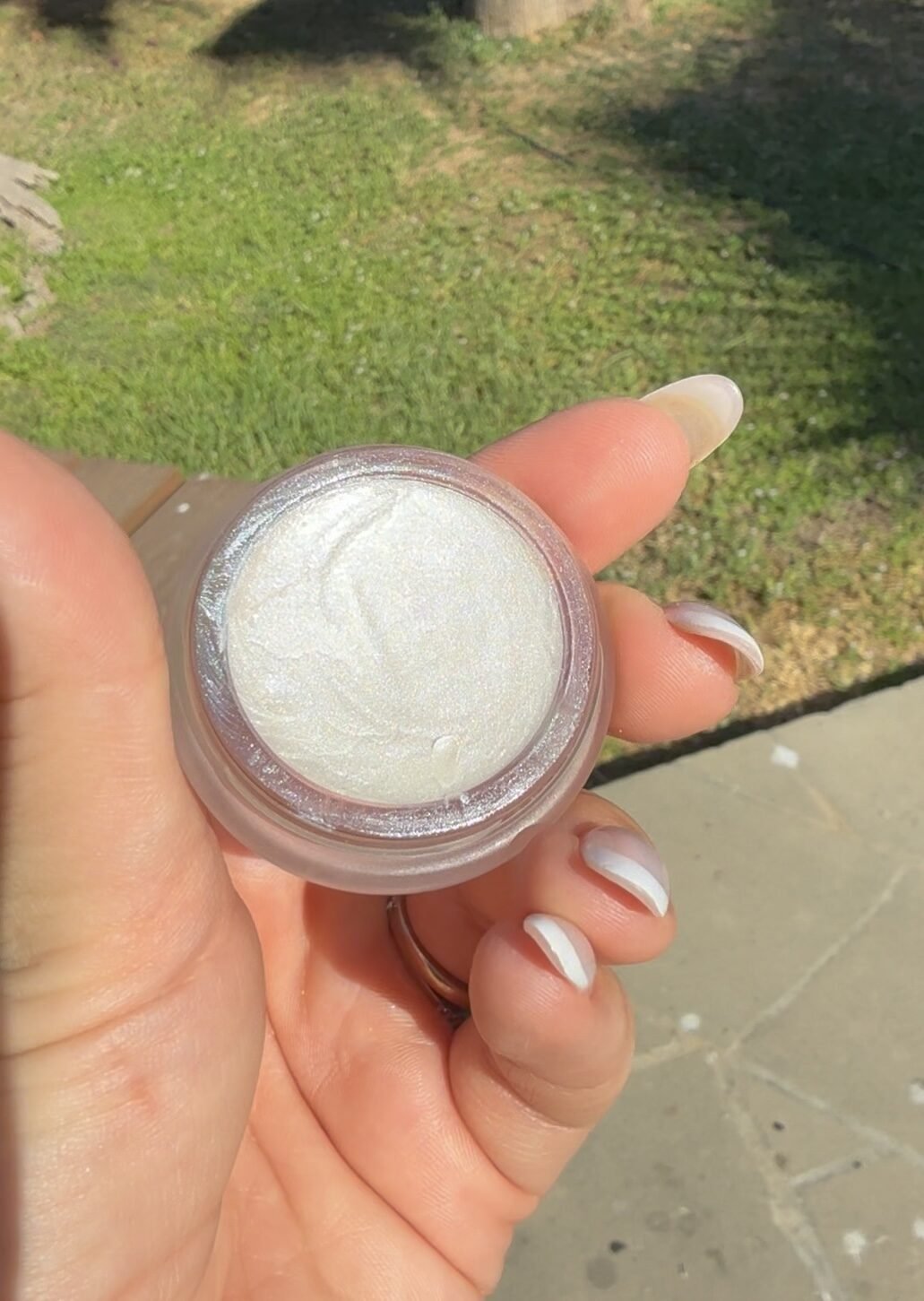 Hand with white manicure holding an open jar of shimmery cream highlighter outdoors on grass.