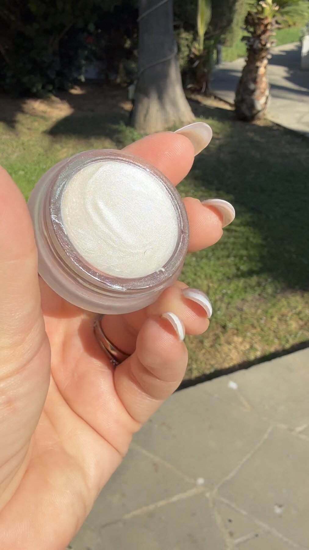 Hand holding an open jar of shimmery white RMS Beauty Living Luminizer cream outdoors on grass.
