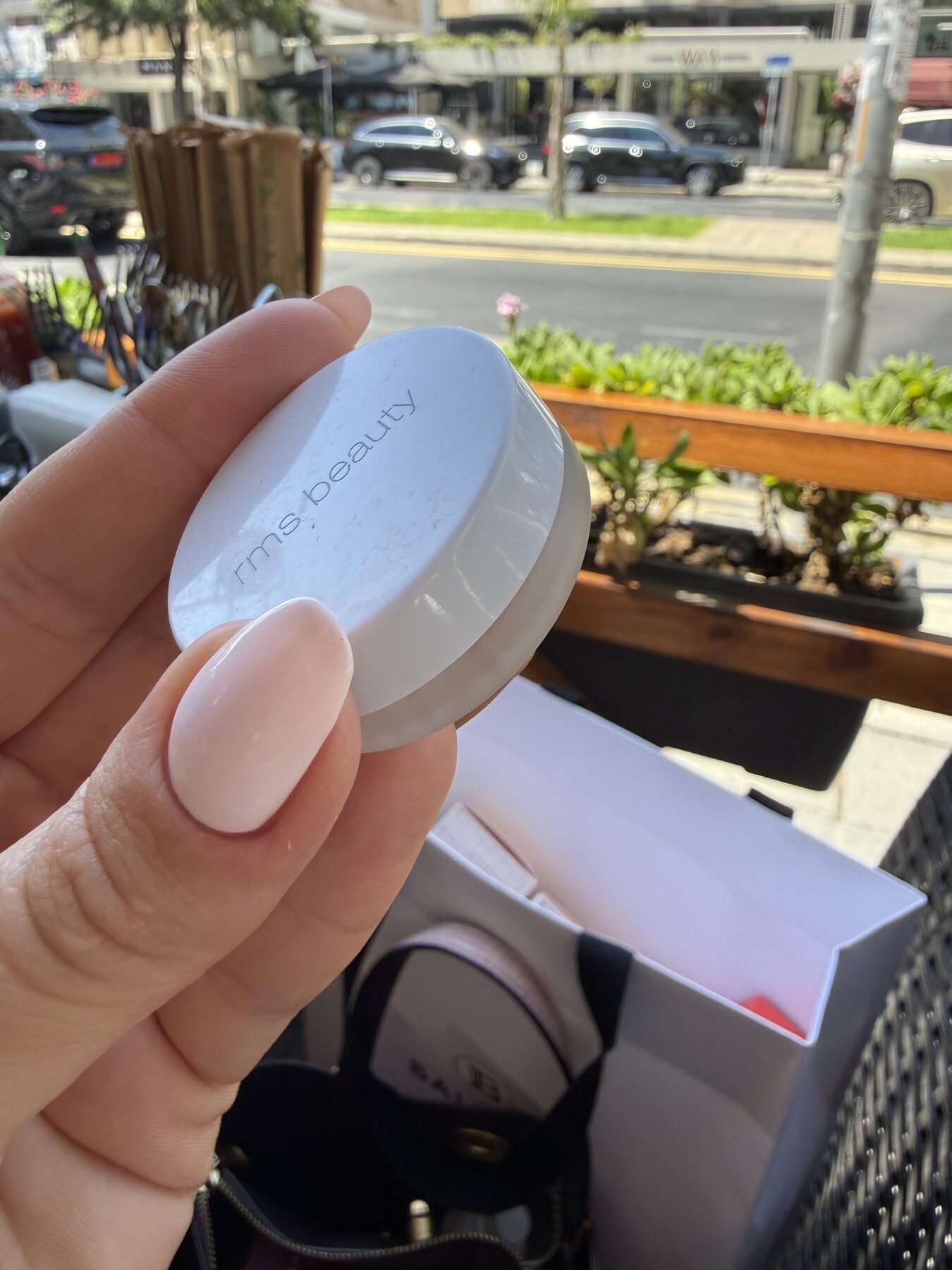 Hand holding a small RMS Beauty jar outdoors, with a street and parked cars in the background.