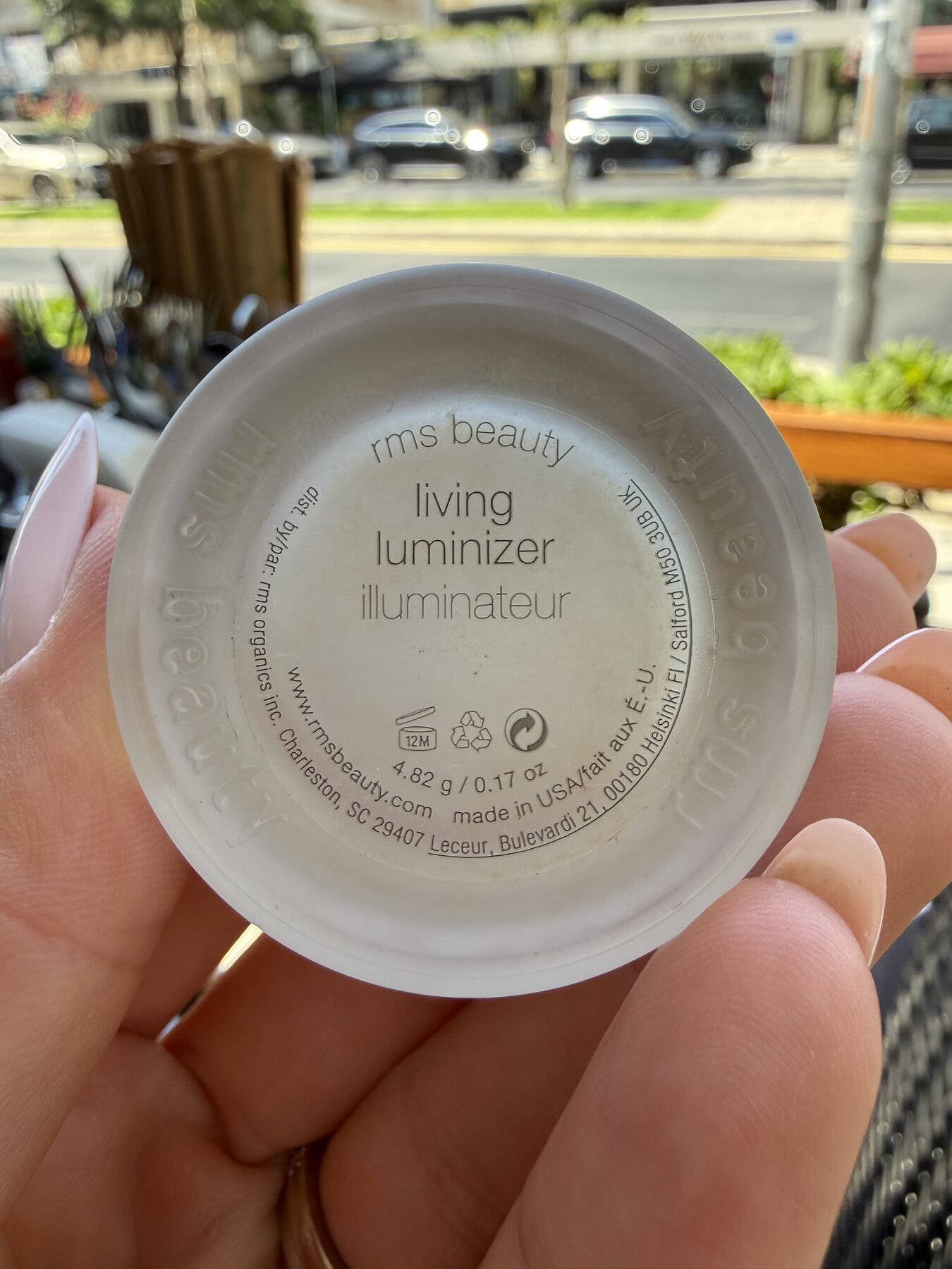 Hand holding the lid of RMS Beauty Living Luminizer, showing engraved product text, on an outdoor street.