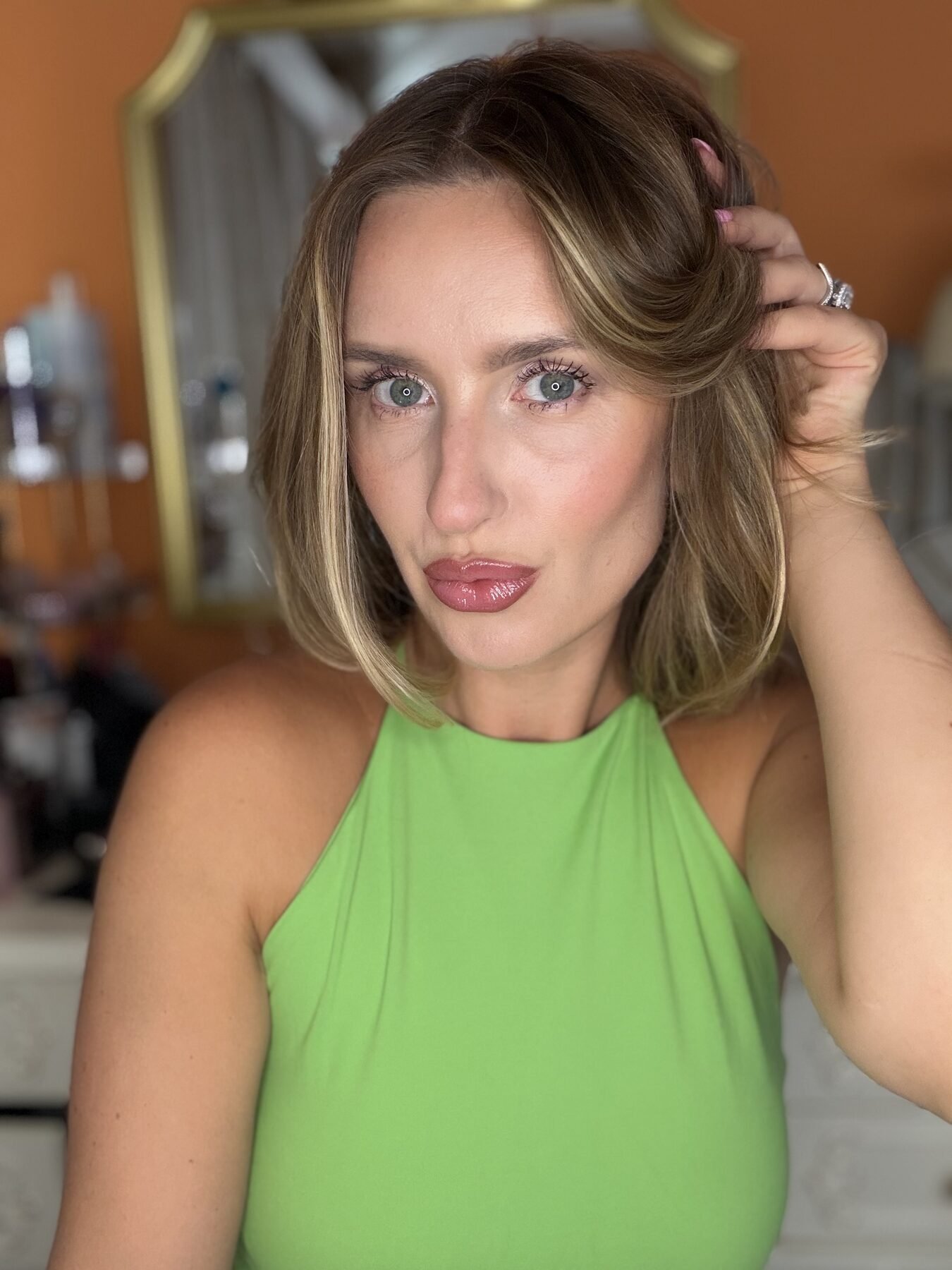Woman in green halter top touches her hair, showing glossy nude-pink lipstick in a mirror selfie.
