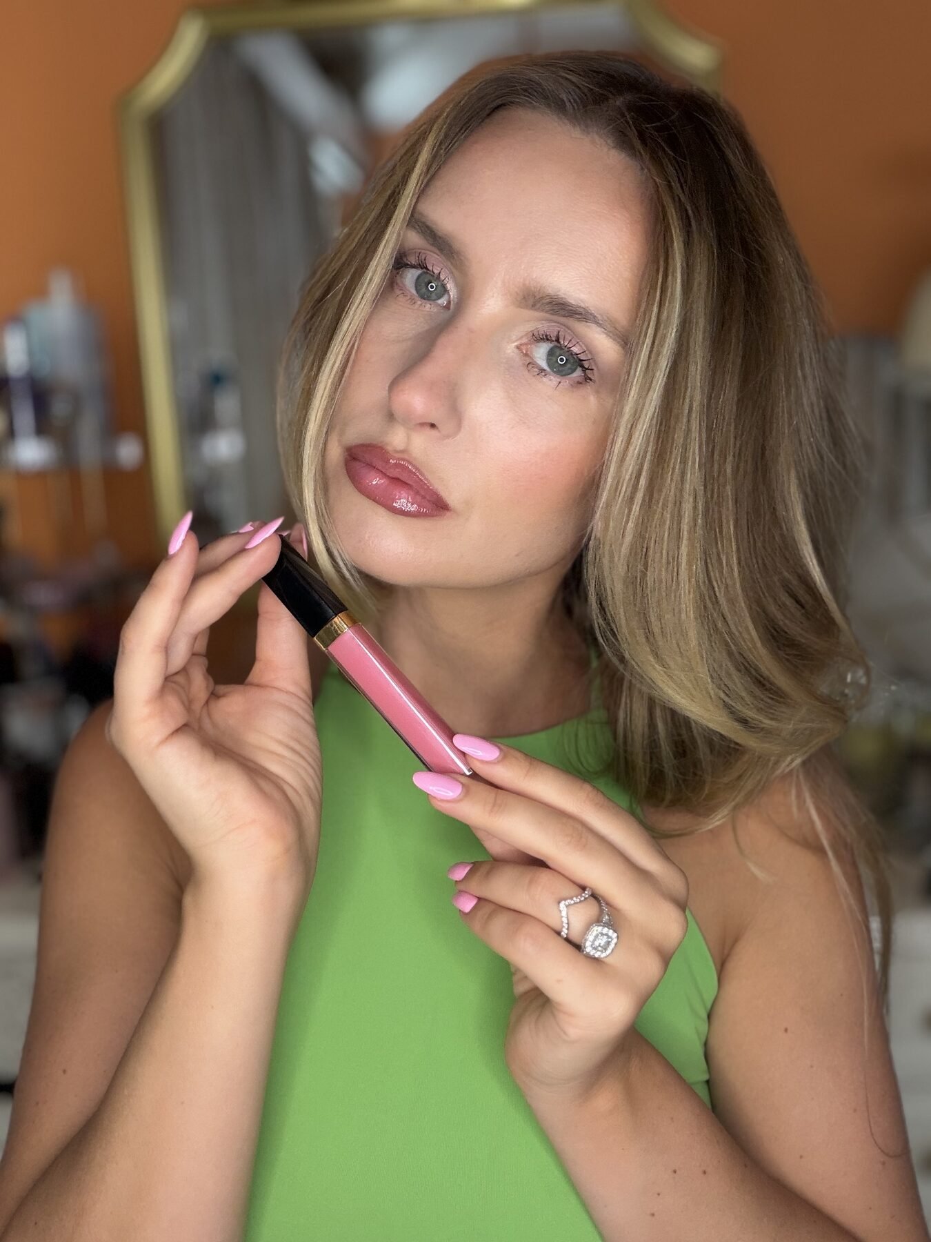 Woman in green top holds open pink lip gloss tube near her glossy tinted lips.