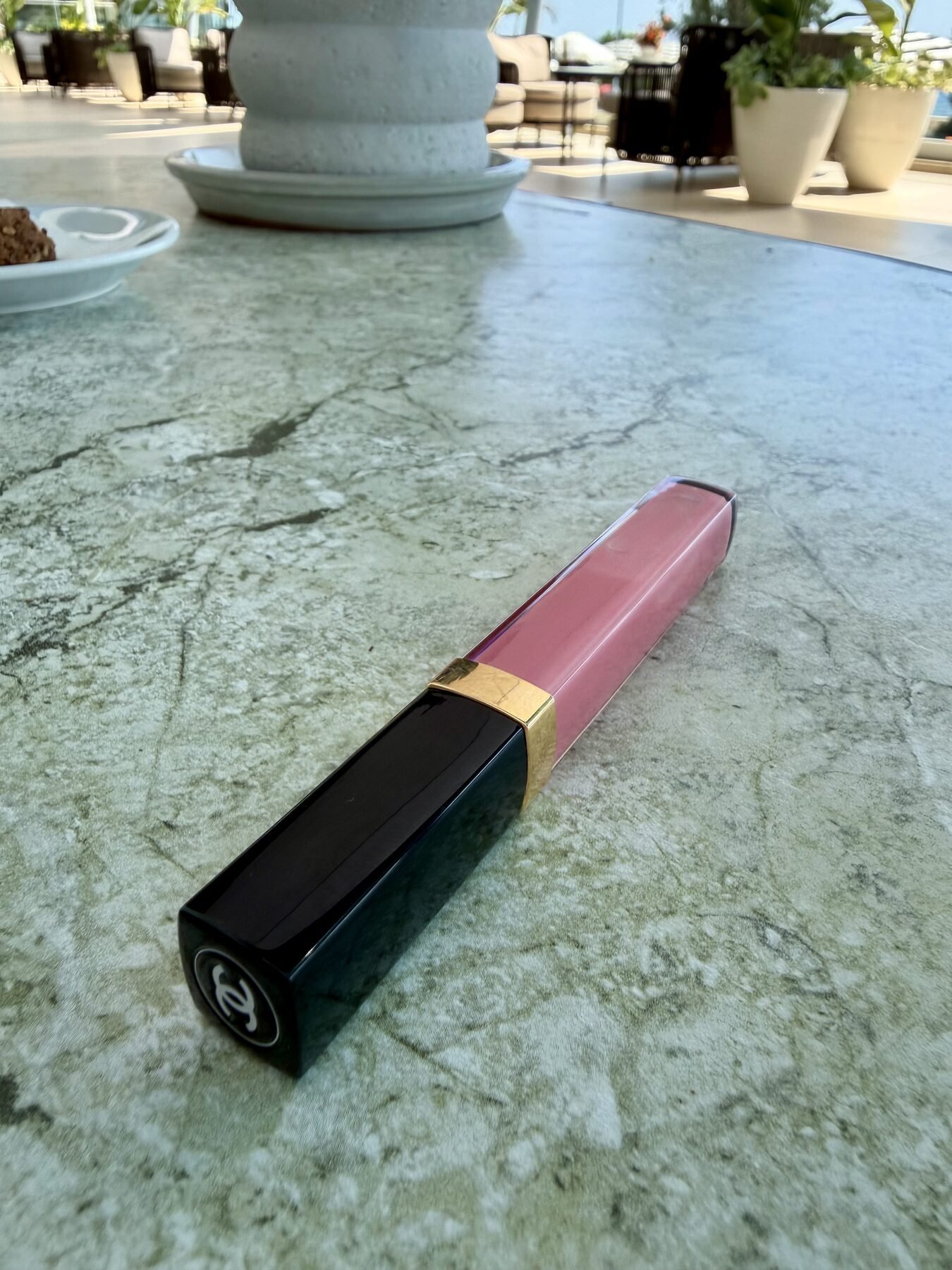 Chanel lip gloss tube lying on a marble table at an outdoor café setting.
