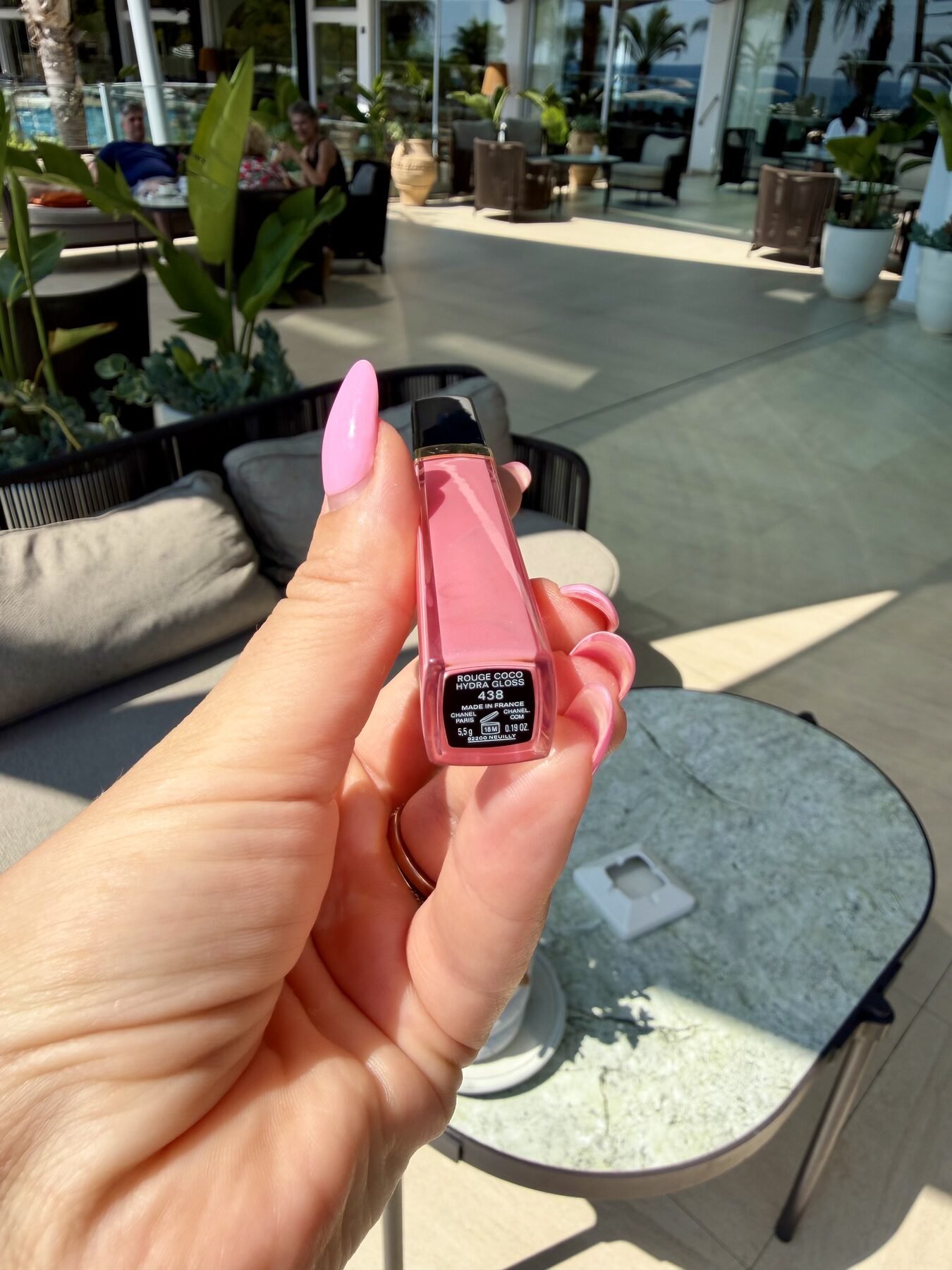 Hand with pink manicure holding Chanel Rouge Coco Hydra Gloss 438 on a sunny outdoor hotel terrace.